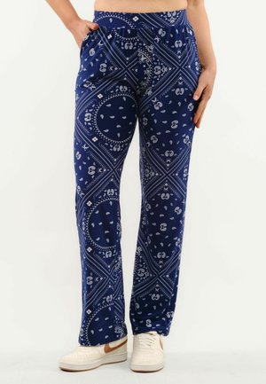 Person iført mørkeblå bukser med hvid paisley-print, elastisk talje, sidelommer og hvide sneakers med brun swoosh.