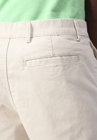 Light beige trousers with a smooth texture, featuring a back pocket and belt loops. Set against a green garment above.