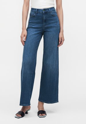 Femme portant un jean bleu taille haute à jambes larges, un haut blanc et des sandales à talons noires ouvertes à l’avant, debout devant un fond uni.
