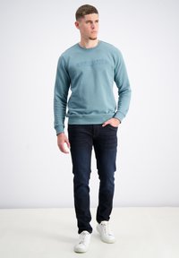 Lichtblauwe sweatshirt met ribbelboorden en zoom, met een textuurlogo. Gedragen met donkere jeans en witte sneakers. Eenvoudige, casual look.