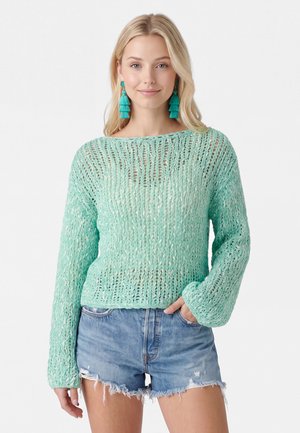 Lyshåret kvinde iført en lysegrøn chunky strikket sweater, blå denimshorts og turkis frynse øreringe, stående med den ene hånd i lommen.