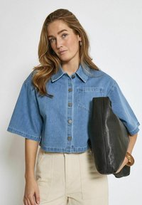 Kurze blaue Jeansjacke mit kurzen Ärmeln, Knopfverschluss und Brusttasche; kombiniert mit einer schwarzen Lederhandtasche.