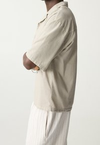 Personne à la peau foncée portant une chemise beige à manches courtes et un pantalon à rayures de couleur claire, se tenant les bras croisés devant un fond uni.