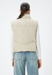 Beige Steppweste mit hohem Kragen und Chevron-Steppung, über einem hellen Langarmshirt getragen und mit hellblauen Jeans kombiniert.