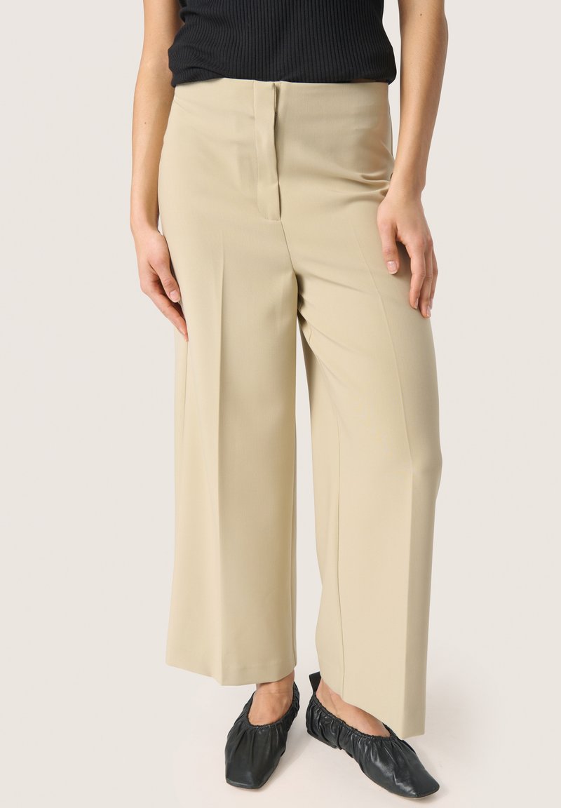 Beige bredde bukser med en glat tekstur, der har en forfure og lynlås lukning. Sammen med sorte slip-on sko.