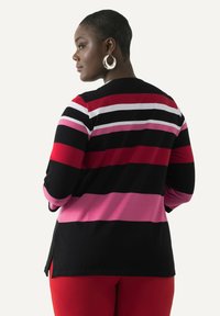 Schwarze Strickjacke mit horizontalen Streifen in Rot, Pink und Weiß. Verfügt über lange Ärmel und seitliche Schlitze. Unterhalb sind rote, eng anliegende Hose sichtbar.
