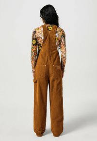 Brown corduroy overalls with a relaxed fit, featuring adjustable straps, front pockets, and a textured finish. Colorful patterned long-sleeve top underneath.