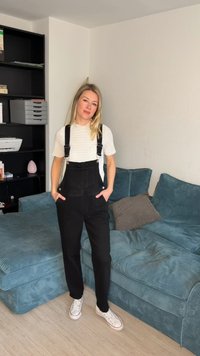 Kvinde med blondt hår iført hvid strikbluse og sorte overalls står med hænderne i lommerne i en stue ved siden af en tælsfarvet sofa.