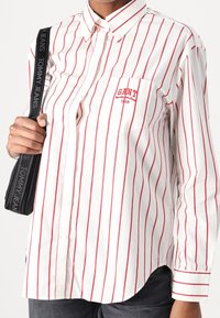 Camicia bianca con bottoni e righe verticali rosse, dotata di tasca sul petto con logo "GANT", e accompagnata da una tracolla nera.