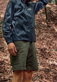 Person in einer marineblauen Windjacke und olivgrünen Cargo-Shorts, der in einem Wald mit fallenem Laub auf dem Boden steht.