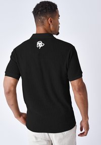 Camiseta polo negra con canales y mangas cortas. Presenta un cuello doblado y un logo bordado en la parte posterior, mostrado desde la vista trasera.