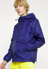 Giacca viola puff con cappuccio, caratterizzata da una texture trapuntata, tasca con zip frontale e polsini elastici. Abbinata a pantaloni shorts gialli.