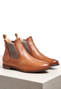 Stivali ankle in pelle marrone con pannelli laterali elasticizzati, caratterizzati da perforazioni decorative, punta rotonda e suola piatta in gomma. Texture liscia.