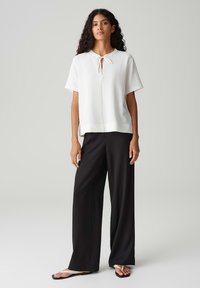 Haut blanc à manches courtes avec un décolleté en trou de serrure et un lien, associé à un pantalon large noir. Le tissu a une finition texturée.