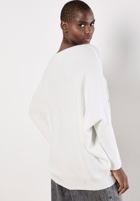 Biały, oversize'owy sweter dzianinowy z opadającymi rękawami, prążkowaną strukturą i szerokim dekoltem, w zestawie z szarymi dżinsami.