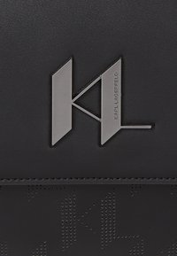 Pelle nera текстurizzata con motivi in rilievo. Logo in metallo argentato "KL" con incisione "KARL LAGERFELD". Design pulito e moderno con linee nette.