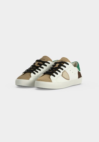 Paire de baskets basses avec empiècements beige, marron et blanc, lacets noirs, semelles blanches et touches de vert sur l’arrière du talon.