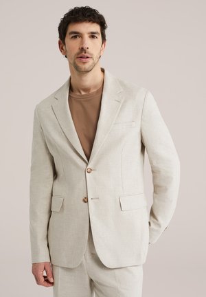 Homme portant une veste de costume beige clair et un pantalon assorti par-dessus un t-shirt marron à col rond, debout devant un fond uni clair.