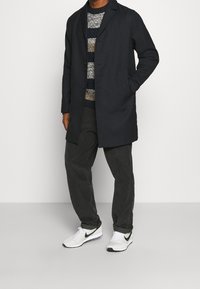 Tmavě modrý kabát s přizpůsobeným střihem, nošený přes pruhovaný svetr, spárovaný se šedými manšestrovými kalhotami a bílými teniskami s černým logem Swoosh.