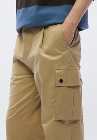 Beežid tööpüksid küljetaskuga, millel on mustad nupud. Valmistatud siledast ja kergest kangast, millel on voldid lisatugevuse jaoks.