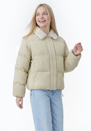 Jeune femme souriante, portant une doudoune beige avec un col en polaire blanc et un jean bleu clair, debout devant un fond blanc.
