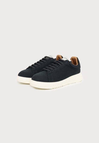 Paire de baskets basses noires avec semelles épaisses blanches et doublure intérieure marron, présentées sur un fond gris clair.
