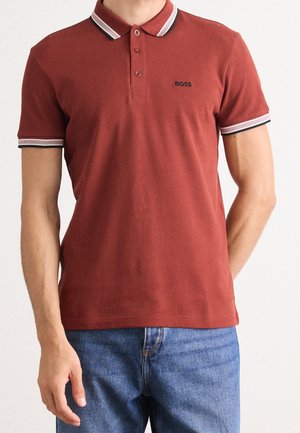 Mand iført en rustfarvet polo med hvide og sorte striber på krave og ærmekanter, kombineret med blå jeans.