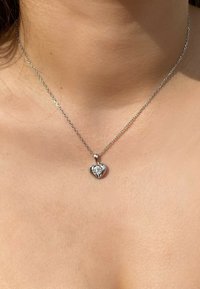 Collier en chaîne argentée avec un pendentif en forme de cœur, orné d'une pierre précieuse claire au centre, reflétant la lumière sur un fond de teint.