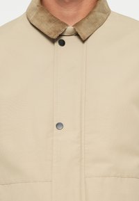 Beige outerwear jacket featuring a corduroy collar, snap closure, and smooth texture with visible stitching and a clean design.