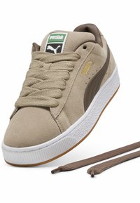 Beige suede sneaker with cream rubber sole, brown accents, and flat laces. Features a green and white logo tag and gold branding on the side.