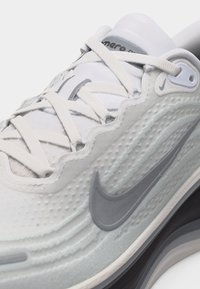 Κοντινό πλάνο λευκού αθλητικού παπουτσιού Nike με υφή στην επάνω επιφάνεια, γκρι λογότυπο swoosh, λευκά κορδόνια και ορατή επενδεδυμένη σόλα.