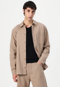 Beige gerippte Knopfleiste mit einer Brusttasche, kombiniert mit einem schwarzen Rundhalsausschnitt-Shirt und passenden beige Hosen. Glatte Textur.