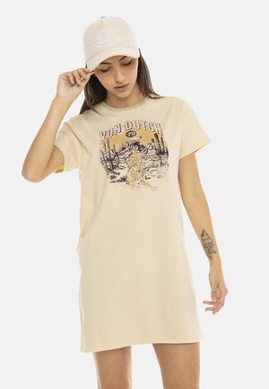 Femme portant une casquette beige Von Dutch et une robe t-shirt oversize assortie avec un graphisme désert et tigre, tenant la visière de la casquette de la main gauche.