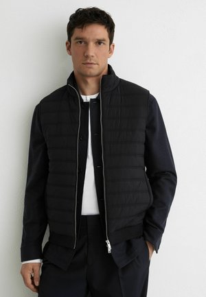 Mand iført en sort quiltet vest med lynlås over en marineblå skjorte og hvid undertrøje, stående mod en ensfarvet hvid væg.