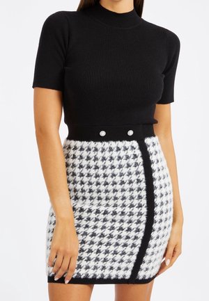 Femme portant un haut court à manches courtes en tissu côtelé noir, rentré dans une mini-jupe à motif pied-de-poule noir et blanc avec deux boutons décoratifs.