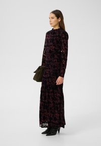 Langärmliges, dunkles, florales Samtkleid mit hohem Ausschnitt, gerafftem Oberteil und gestuftem Rock, kombiniert mit einer dunkelgrünen Clutch und spitzen Absätzen.
