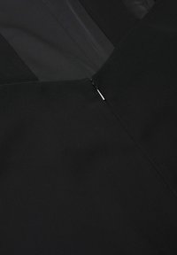 Gros plan sur un vêtement noir présentant une fermeture éclair partiellement visible avec une languette argentée sur un tissu texturé.