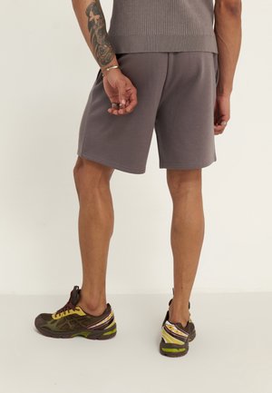 Mężczyzna w szarych szortach, brązowo-żółtych tenisówkach i bezrękawniku, pokazujący wytatuowane lewe przedramię i pierścienie na dłoniach.