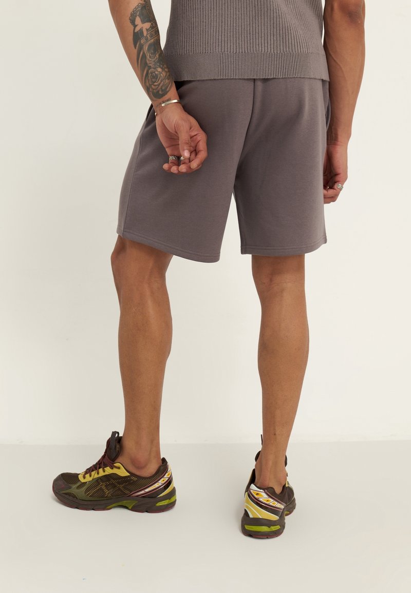 Homme portant un short gris, des baskets marron et jaunes, et un débardeur, montrant son avant-bras gauche tatoué et des bagues aux doigts.