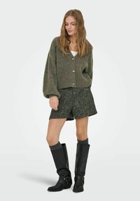 Cardigan verde lavorato a maglia con polsini a coste, pantaloni corti a righe nere e bianche, calze grigie fino al ginocchio e stivali di pelle nera con fibbie.