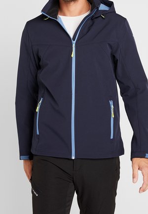 Mężczyzna ubrany w granatową kurtkę z kapturem z jasnoniebieskim zamkiem błyskawicznym i żółtymi uchwytami zamka, w połączeniu z czarnymi spodniami.