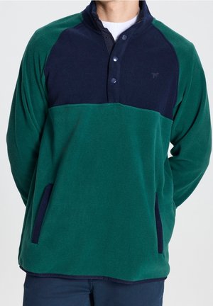 Homme portant un pull polaire bicolore vert et marine avec boutons-pression et poches latérales, associé à un pantalon foncé.