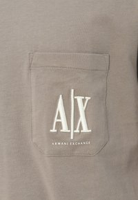 T-shirt grigio con tessuto testurizzato. Presenta una tasca sul petto sinistro con logo "A|X" ricamato in bianco e il testo "ARMANI EXCHANGE" sottostante.