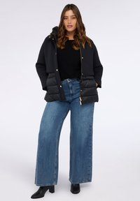 Mustat puffaliivit, joissa on vetoketju- ja napitus, huppu ja erottuva neulekangas. Yhdistetty leveleginsien kanssa sinisissä farkuissa ja mustissa saappaissa.