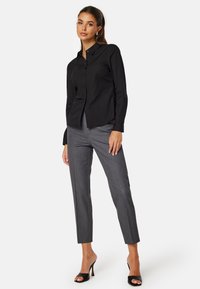 Camicia nera con bottoni realizzata in tessuto liscio, abbinata a pantaloni grigi su misura con sottili righe. Indossata con sandali neri con tacco.