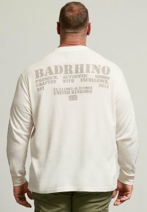 Mand med kort hår iført en hvid langærmet skjorte med "BADRHINO" og tekst på bagsiden, kombineret med olivengrønne bukser.