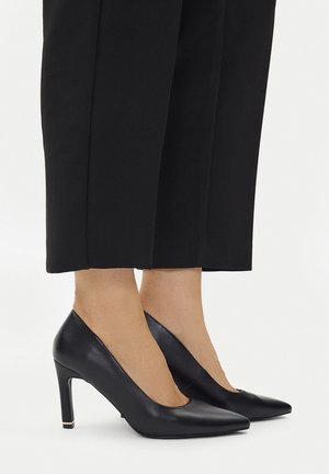 Czarne szpilki wykonane z gładkiej skóry, z ostrym noskiem i smukłym obcasem z złotym akcentem. Połączone z czarnymi spodniami.