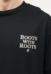 Μαύρο πουκάμισο με λευκό κείμενο «BOOTS WITH ROOTS» και ένα μικρό λογότυπο δέντρου κάτω από το κείμενο στην περιοχή του στήθους.