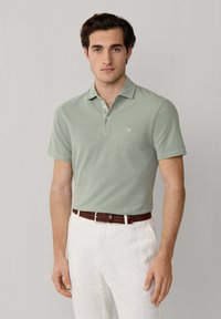 Jeune homme portant un polo à manches courtes vert clair, un pantalon blanc et une ceinture marron, debout devant un fond clair uni.