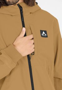 Brun vattentät jacka med framdragkedja, justerbar huva och bröstficka med en svart logotypetikett. Slät tygtextur.